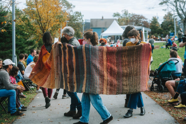A group of people wearing masks walk in a circle down the path of QUIET PARADE, carrying a three-foot wide, very long, multicolored, striped, hand-woven blanket.