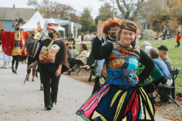 A person wearing a colorful patterned dress waves at the crowd while leading their group of models down the path at QUIET PARADE.