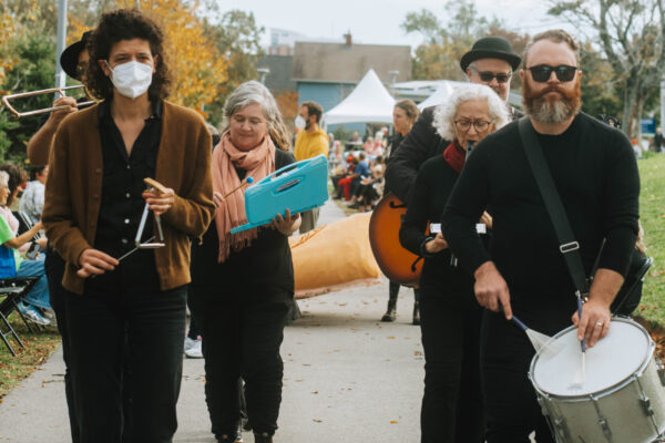 The Hush Band, six musicians wearing dark clothes, softly play their instruments in QUIET PARADE. They are arranged in rows of two, with percussionists in the front, wind instruments in the centre, and a guitarist and glockenspielist in the back row.