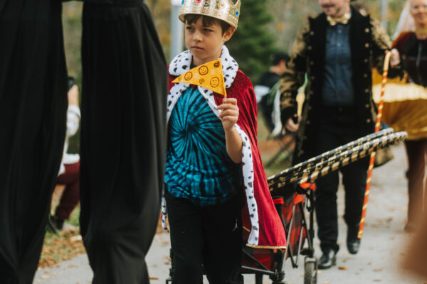A group of people in circus costumes walk down a paved path in a line. The image is focused on a young child wearing a crown and red cape, waving a small yellow flag and dragging a wagon full of hula hoops. In front of them is a person visible from the waist down and is dressed in black and walking on stilts. Walking behind the child and out of focus, is a person in a top hat and long coat.
