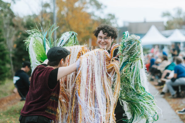 Two people laugh and wave their arms at one another during QUIET PARADE. They each have long, voluminous, stringed wearable art on their arms that resemble wings made of yarn. One person is wearing orange, white, and purple yarn and has their back to the camera and the other person is wearing green, white, and blue yarn