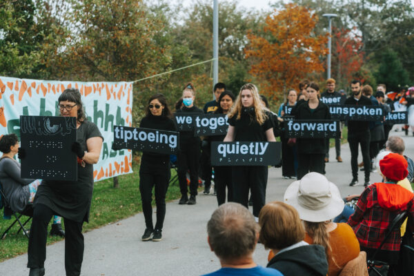 A group of over 10 individuals wearing black; each one is walking and carrying a black sign with white lettering and braille that forms a moving poem at QUIET PARADE.