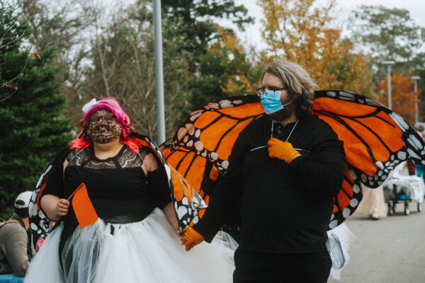 Two people wearing orange monarch butterfly wings and black shirts smile and hold hands. The person on the left has pink hair, a floral face mask, and white tutu. The person on the right has brown hair, a blue medical mask, and orange gloves.