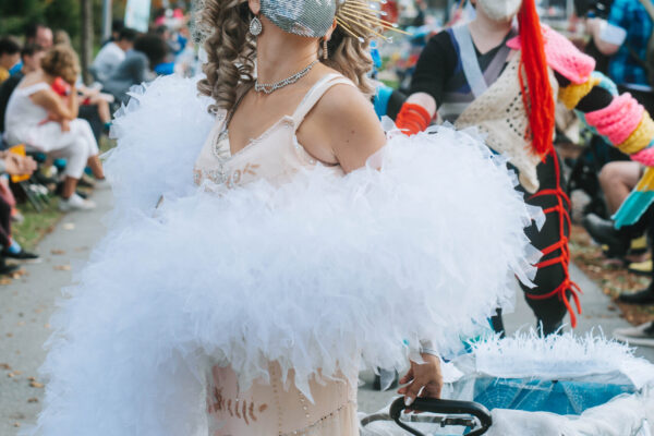 A burlesque performer wearing an extravagant outfit smiles at the camera beneath a silver mask. They have an ornate gold crown on their head, curly blond hair, silver makeup, and an extra-large feather boa that wraps around their body and peach-colored gown. They are walking and pulling a wagon along the parade route at QUIET PARADE.