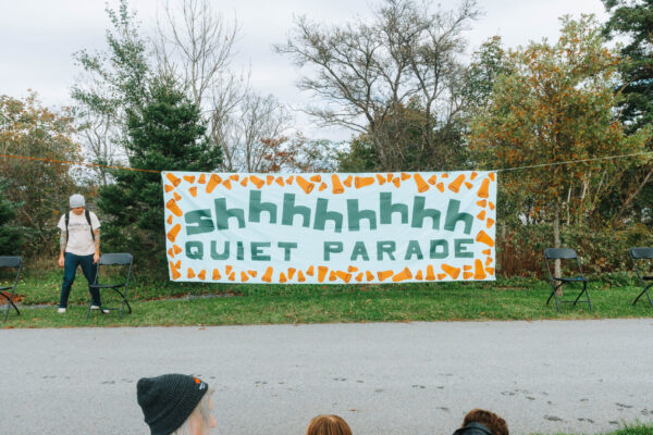 Three parade spectators sit on the grass at the edge of a path with their backs to the camera, facing a banner on the other side of the path. The banner says “shhhhhhhh” on top and “QUIET PARADE” on the bottom in green letters. The text is bordered by illustrations of orange foam earplugs.