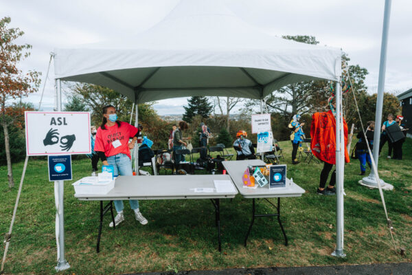 A MSVU Art Gallery staff member stands at the QUIET PARADE Welcome Table. They are standing to the left of a table beneath a tent, wearing a bright pink shirt that says “QUIET PARADE,” a nametag, a mask, and blue jeans. There are some QUIET PARADE participants gathered on the grass in background and signage and event supplies on the table in the foreground.
