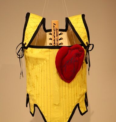 A yellow corset lined with black hanging from the ceiling. The corset has a large red felted heart attached to the chest.